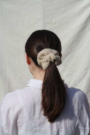 White woman with brown hair wearing facing away from the camera wearing a cream knitted wool scrunchie in her hair. Also wearing a white shirt against a white fabric background.